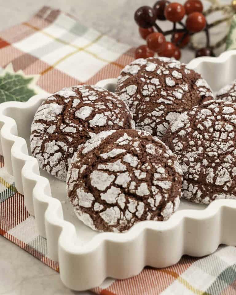Red coloured cookies with cracked outta shell, coated in icing sugar placed in a white dish on christmas decorations in background and red and green checkered tea towel below.