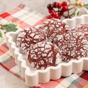 Red coloured cookies with cracked outta shell, coated in icing sugar placed in a white dish on christmas decorations in background and red and green checkered tea towel below.
