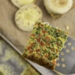 Metal spatula holding a square slice of sheet pan egg above a chopping board with breakfast muffins.