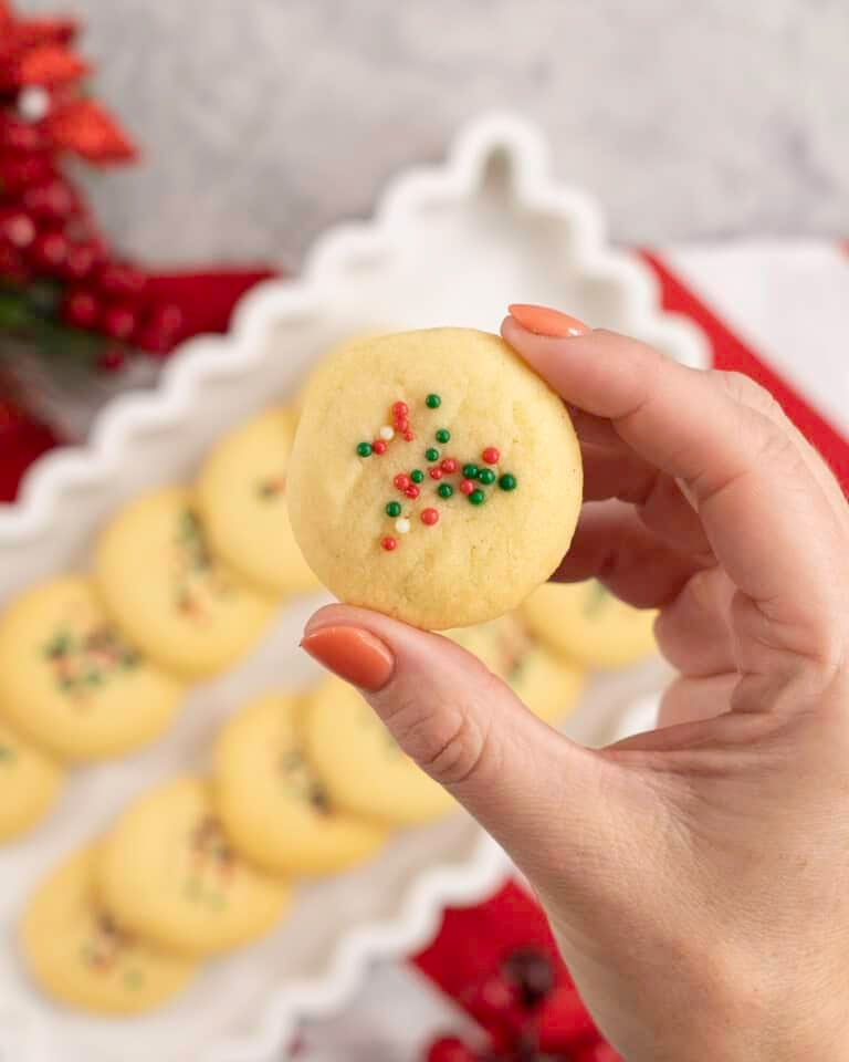 A shortbread cookie decorated with Christmas sprinkles held above a tray of more cookies.